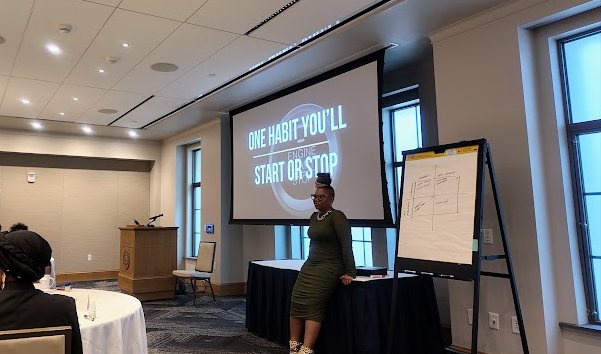 Rhonda Gipson-Willis stands beside a projected slide reading One Habit
                You'll Start or Stop, next to a flip chart with a hand drawn grid, while
                a participant sits at a round banquet table in the foreground.