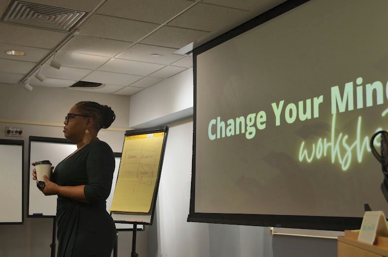 Rhonda Gipson-Willis stands in profile holding a coffee cup, mid
                    thought, beside a flip chart and a projected slide reading Change
                    Your Mind workshop.