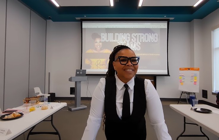 Rhonda Gipson-Willis smiles toward the camera in a training room, wearing
                a white shirt and black waistcoat, with a Building Strong Teams slide on
                the screen behind her.