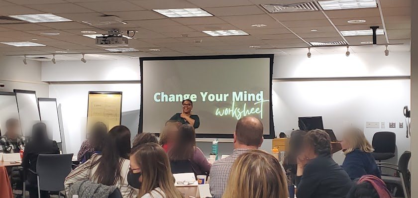 Around fifteen people seated at tables in a training room, facing a
                projected slide that reads Change Your Mind worksheet, while Rhonda
                Gipson-Willis speaks from the front of the room.
